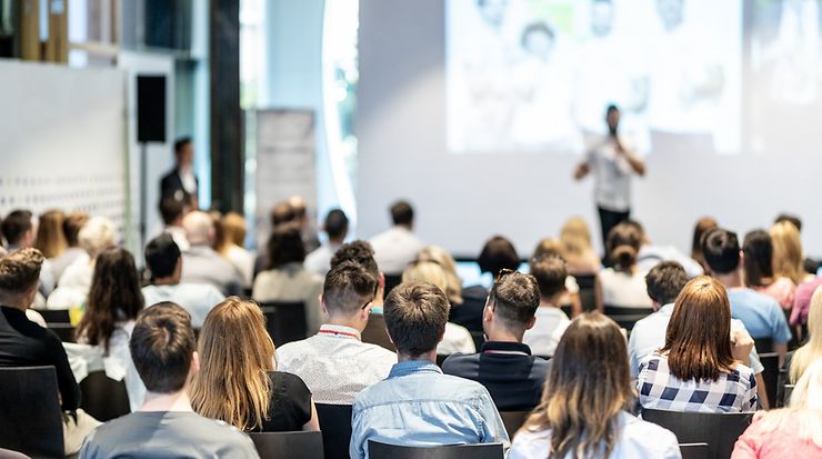 Publikum, das auf eine Bühne guckt, wo eine Person an einem Mikrofon steht