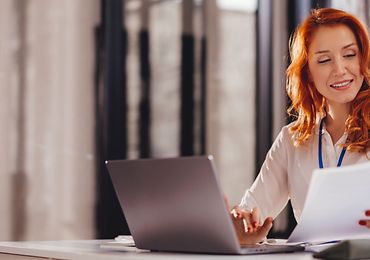 Frau sitzt am Laptop und hat ein Stapel Papier in der Hand