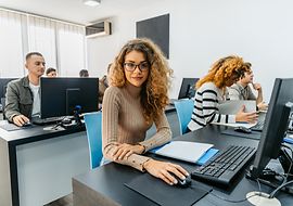 In einem Schulungsraum sitzen mehrere Personen an Computern. Im Vordergrund - eine weibliche Person schaut in die Kamera.