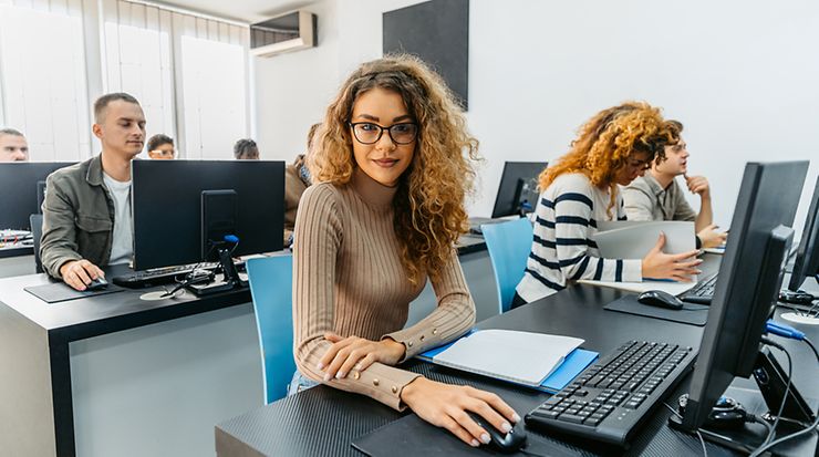 In einem Schulungsraum sitzen mehrere Personen an Computern. Im Vordergrund - eine weibliche Person schaut in die Kamera.