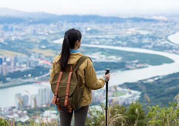 Frau mit Rucksack und Wanderstock steht vor einer Stadt und schaut auf diese