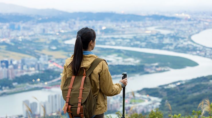 Frau mit Rucksack und Wanderstock steht vor einer Stadt und schaut auf diese