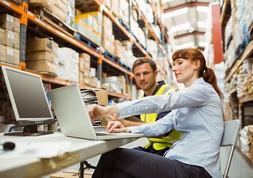 Zwei Personen arbeiten in einem Lager, zeigen auf einen Laptop-Bildschirm und umgeben von Regalen mit Kartons.