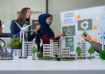 Im Vordergrund eine Modellstadt mit Windrad auf einem Tisch, im Hintergrund zwei Personen an einem Whiteboard