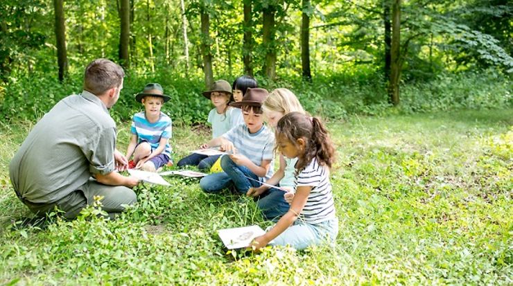 Kinder sitzen mit einem Erwachsenen im Wald und lernen aus Büchern