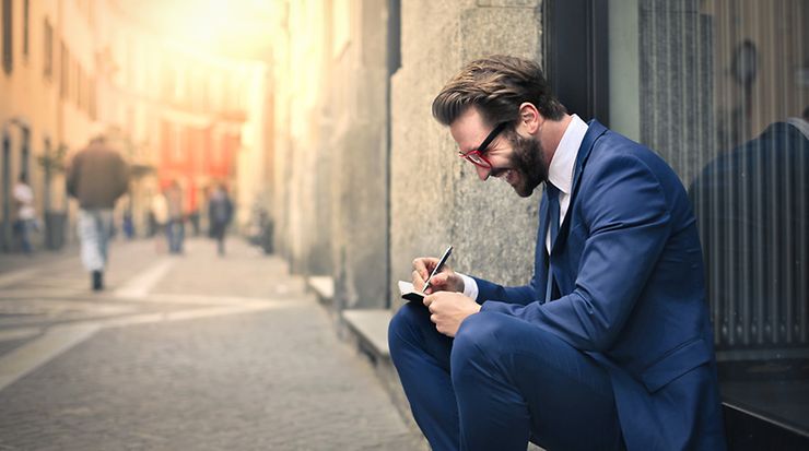 Mann im Anzug sitzt auf Schwelle in einer Straße, schreibt in ein Buch und lächelt