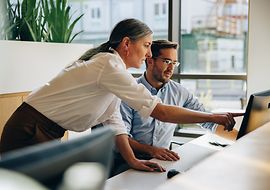 Eine Frau erklärt einem Mann etwas am Arbeitsplatz und zeigt auf einen Computerbildschirm